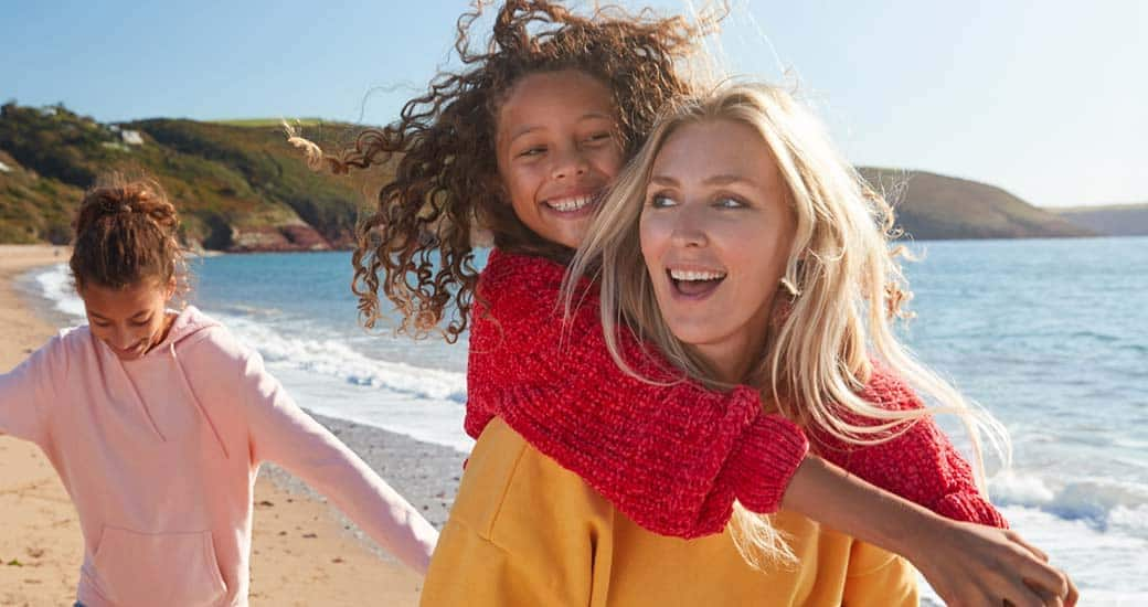 A woman is carrying one of her children on her back. The other one is walking behind them. They are at the beach.