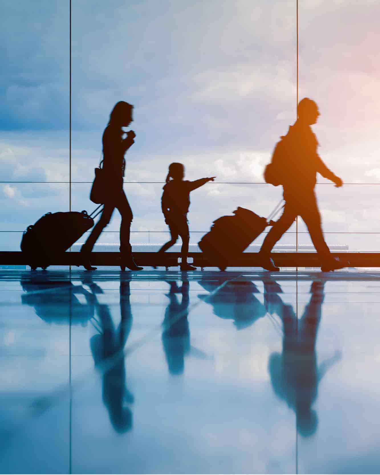 Two parents and a child silhouette are walking through an airport in front of scenic windows. They are carrying luggage. 
