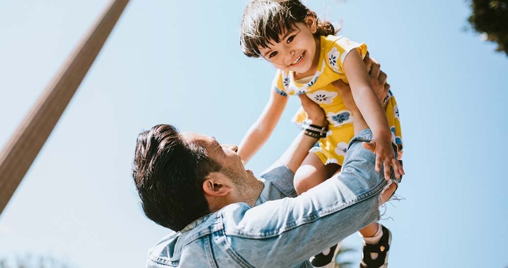 Father lifts his young daughter into the air. The young girl is happy. 